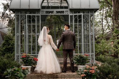 back view of bride and groom in safety masks holding hands near alcove in park