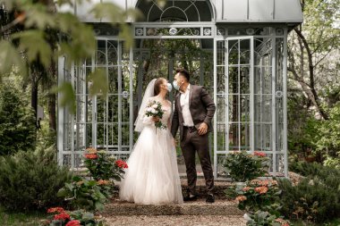 full length view of young newlyweds in medical masks kissing near alcove in park