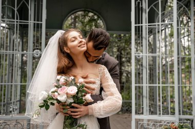 young man kissing neck of happy bride holding wedding bouquet in park