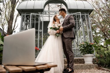 young newlyweds in medical masks kissing outdoors near laptop on blurred foreground