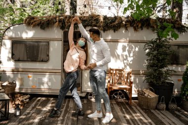 young couple in protective masks dancing near camper in forest