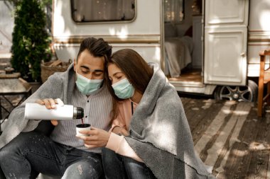 couple in medical masks pouring warm coffee while sitting under blanket near camper