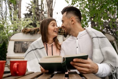 cheerful couple looking at each other while sitting with book in camping