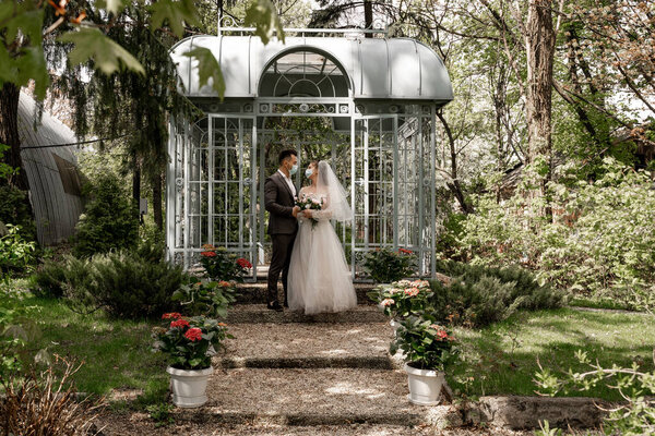 full length view of newlyweds in medical masks standing near alcove in park