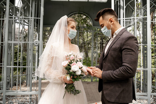 young man in medical mask putting wedding ring on finger of bride