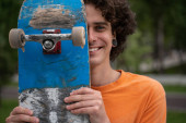 fröhlicher Mann blickt in Kamera, während er sein Gesicht mit Skateboard verdeckt