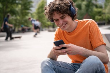 Kulaklıklı mutlu genç adam kaykay parkında cep telefonuyla sohbet ediyor.