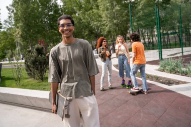 Genç ve neşeli Afrikalı Amerikalı adam arka planda arkadaşlarının yanında paten tutuyor.