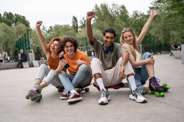 excited multicultural skaters showing win gesture while sitting on road in skate park