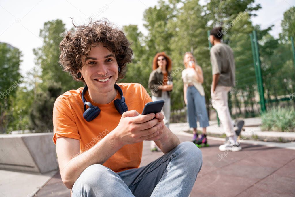 curly man with mobile phone smiling at camera near blurred multiethnic skaters