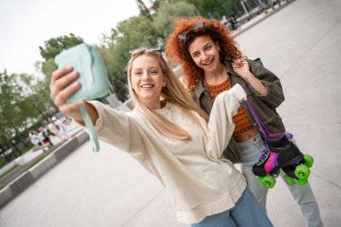 Patenli kıvırcık kadın, arkadaşının yanında gülümseyerek dijital kamerada selfie çekiyor.