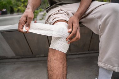 Afrika kökenli Amerikalı bir adam bankta oturuyor ve yaralı dizini bandajlıyor.