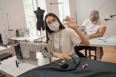 Tıbbi maskeli Asyalı terzi dikiş makinesi ve iş arkadaşının yanında el sallıyor. 