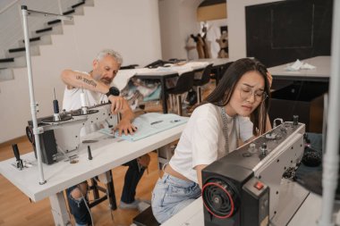 Yorgun Asyalı terzi dikiş makinesi ve iş arkadaşının yanında oturuyor. 