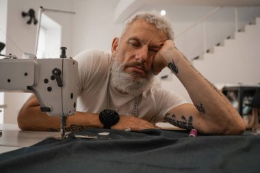 Yorgun terzi kumaş ve dikiş makinesinin yanında oturuyor. 