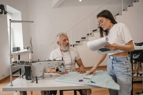 Asiatischer Designer Hält Klemmbrett Neben Kollegin Und Nähmaschine Atelier — Stockfoto