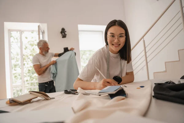 Lächelnder Asiatischer Designer Blickt Während Mit Tuch Arbeitet Die Kamera Stockbild