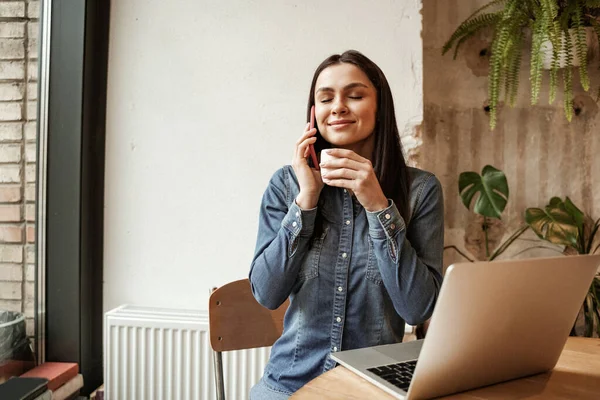 Щаслива жінка тримає чашку кави під час розмови на смартфоні біля ноутбука — стокове фото