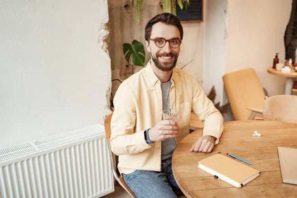 Веселый бородатый мужчина держит стакан воды, сидя рядом с ноутбуком на столе — стоковое фото