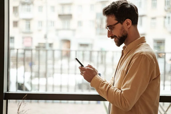Side view of bearded man using smartphone while standing near window — Stock Photo