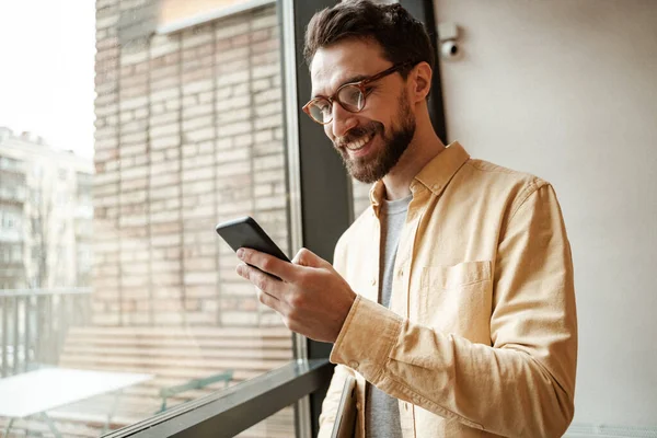 Smiling and bearded man using smartphone while standing near window — Stock Photo