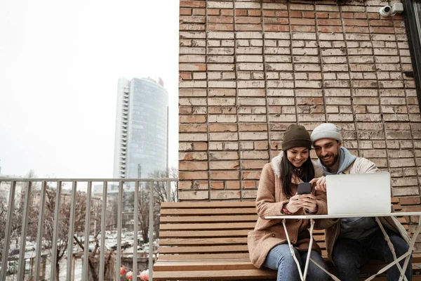 Fröhliche Freiberufler in Mützen schauen auf das Handy neben dem Laptop auf dem Tisch — Stockfoto
