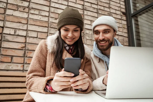 Freelancers felices en gorros de gorro mirando el teléfono inteligente cerca de la computadora portátil en la mesa - foto de stock