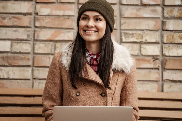 Smiling brunette freelancer in beanie hat using laptop while sitting on bench — Stock Photo