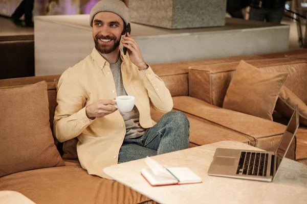 Cheerful bearded man in beanie hat holding cup and talking on smartphone — Stock Photo