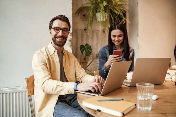 Heureux pigiste dans les lunettes à l'aide d'un ordinateur portable près de la femme dans le café — Photo de stock
