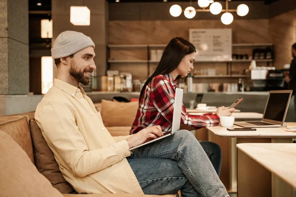 Bearded freelancer in beanie hat typing on laptop near woman on blurred background — Stock Photo