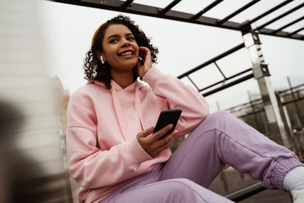 Joyeuse sportive afro-américaine tenant smartphone tout en écoutant de la musique sur le stade en plein air — Photo de stock