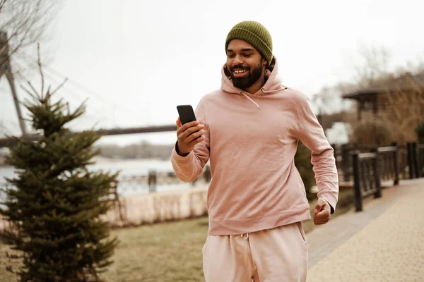 Jogger afro-américain joyeux tenant téléphone portable tout en courant à l'extérieur — Photo de stock