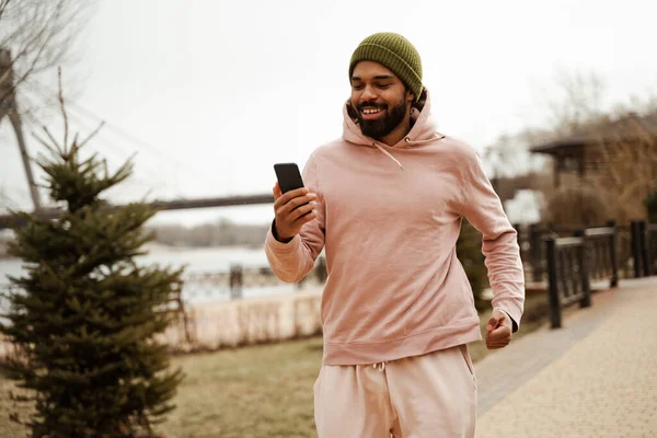 Cheerful african american jogger holding mobile phone while running outdoors — Stock Photo