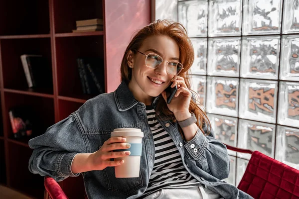 Щасливий рудий студент в окулярах тримає еко-чашку з кавою під час розмови на смартфоні — стокове фото