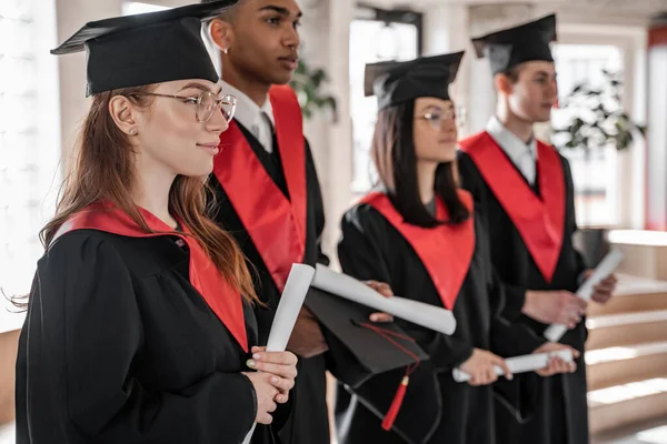 Estudiante feliz cerca de compañeros de clase interracial en vestidos de graduación y gorras con diploma sobre fondo borroso - foto de stock