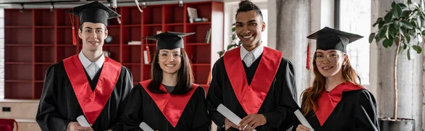 Feliz interracial estudiantes en la graduación vestidos y gorras celebración de diploma, baile 2021, pancarta - foto de stock