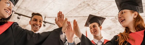 Feliz interracial estudiantes en batas y gorras celebración diploma y dando alta cinco, graduación clase 2021, bandera - foto de stock