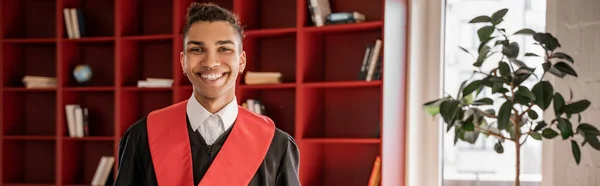 Estudiante afroamericano feliz en vestido de graduación, bandera - foto de stock