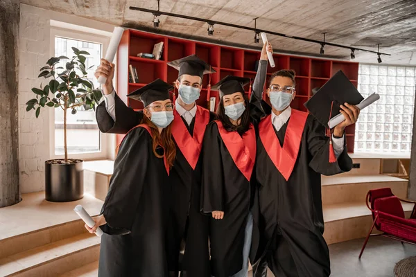 Estudantes multiétnicos em máscaras médicas, vestidos e bonés com diploma, classe de graduação 2021 — Fotografia de Stock