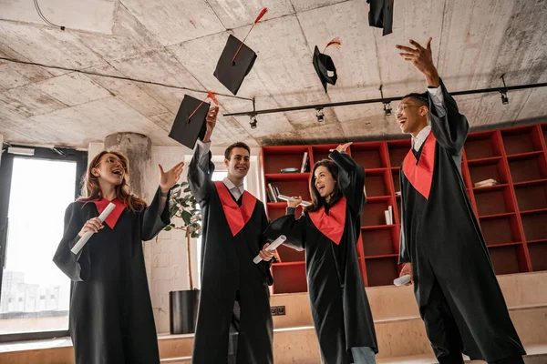 Asombrado interracial estudiantes en batas lanzando gorras y celebración diploma, graduación clase 2021 - foto de stock