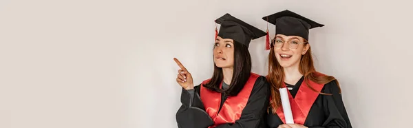 Bastante estudiante en vestido de graduación y gorra señalando con el dedo cerca compañero de clase alegre, senior 2021, pancarta - foto de stock