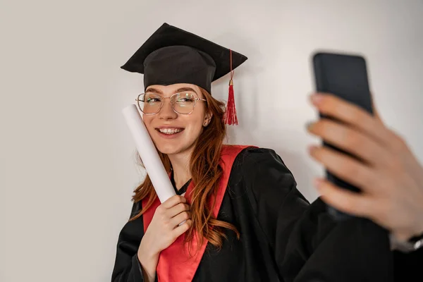 Estudiante alegre en la gorra de graduación y vestido de tomar selfie, senior 2021 - foto de stock