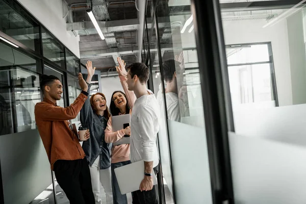 Happy interracial students holding gadgets and coffee to go while giving high five — Stock Photo