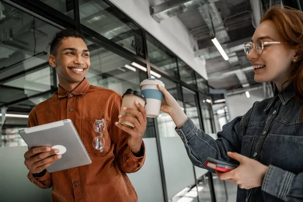 Afrikanischer Student hält digitales Tablet in der Hand und klappert mit fröhlichem Klassenkameraden Pappbecher — Stockfoto