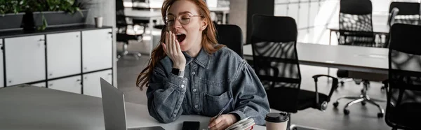 Tired redhead student yawning near gadgets and paper cup on desk, banner — Stock Photo