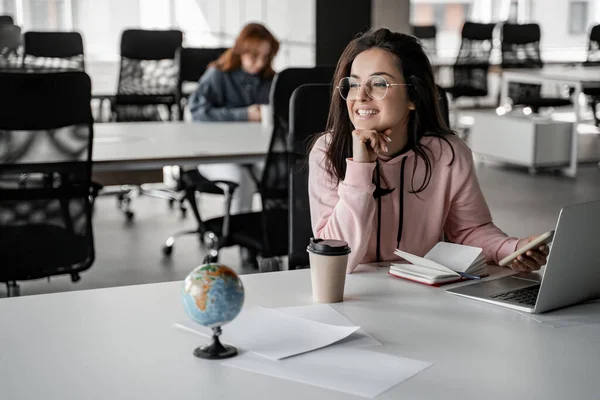 Happy brunette student in glasses looking away while holding smartphone near laptop — Stock Photo