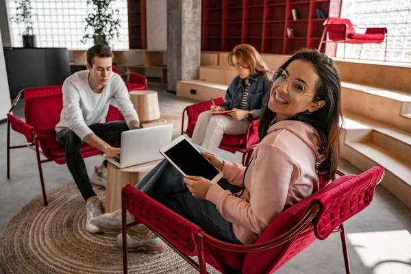 Fröhliche brünette Studentin mit Brille hält digitales Tablet mit leerem Bildschirm in der Nähe von Klassenkameraden auf verschwommenem Hintergrund — Stockfoto