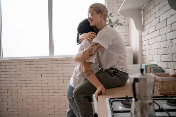 Tattooed woman in glasses sitting on kitchen table and hugging brunette girlfriend — Stock Photo