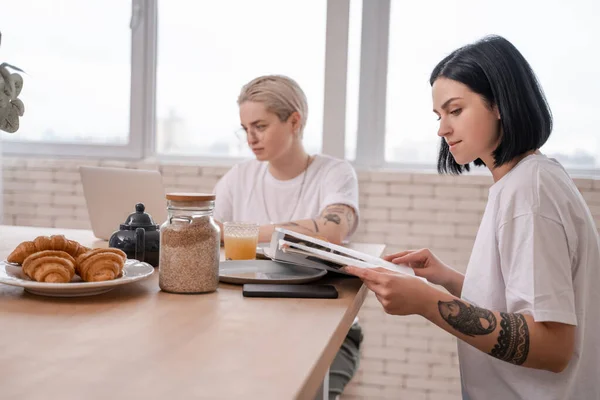 Brunette woman reading magazine near girlfriend using laptop on blurred background — Stock Photo